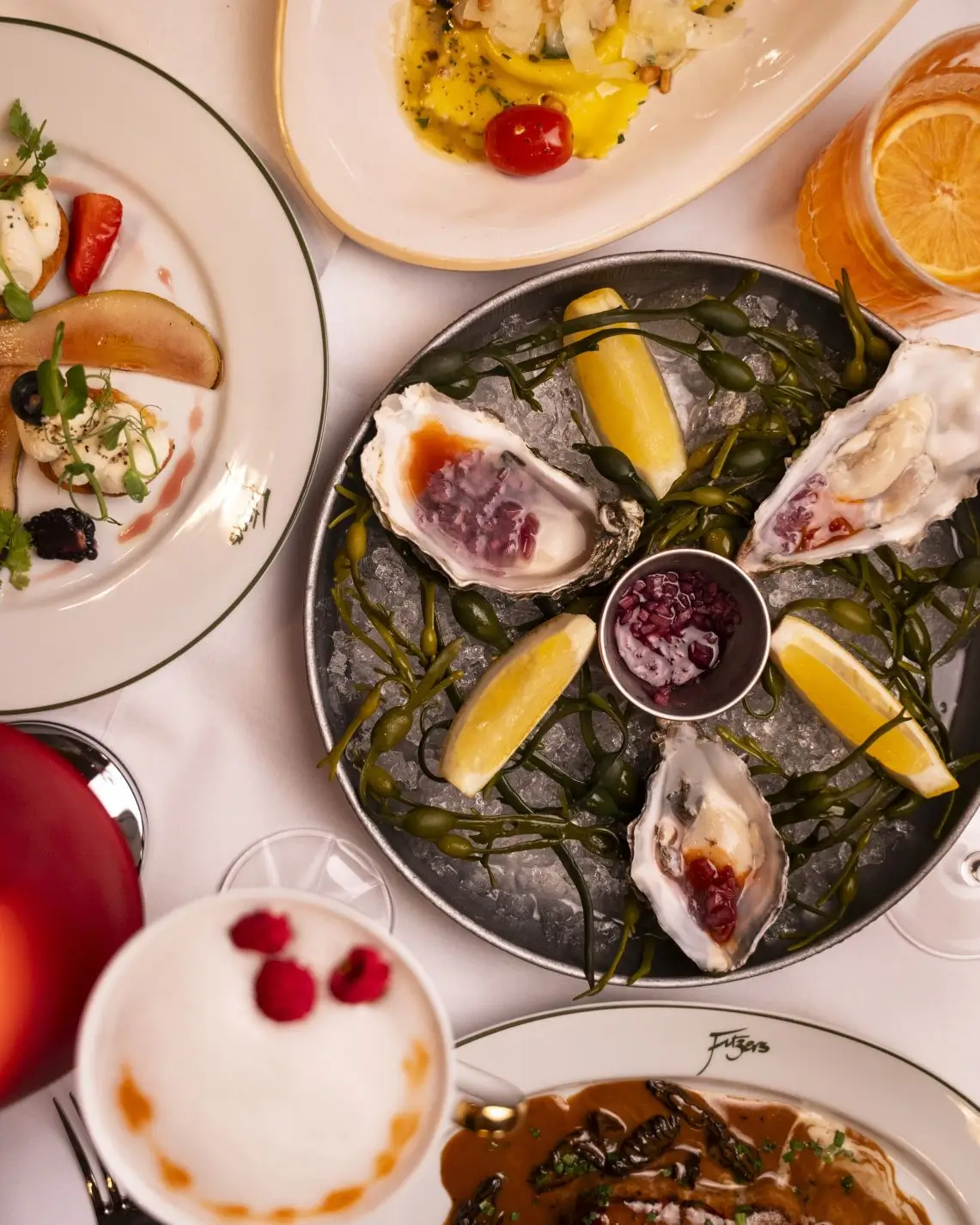 A top-down view of a table at Fitzers Bistro, laden with dishes including fresh oysters, goat cheese crostini, pasta ravioli, and cocktails.